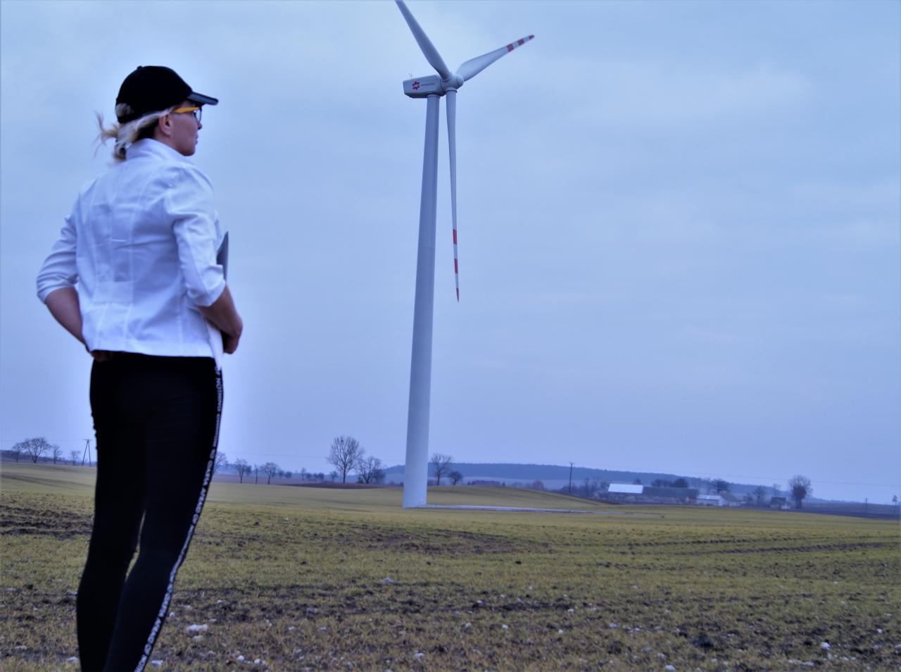 Kobieta w czapce i białej kurtce stoi na polu z wiatrakiem w tle, patrząc w dal. Krajobraz wiejski, pochmurne niebo.