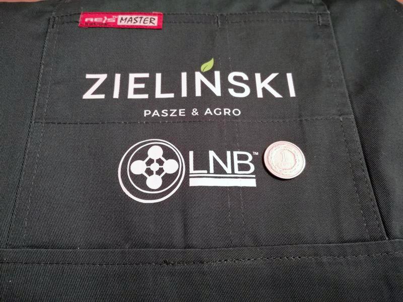Czarny fartuch roboczy z nadrukowanym białym logo firmy ZIELIŃSKI PASZE & AGRO oraz logiem LNB, obok którego leży moneta jednozłotowa dla skali.