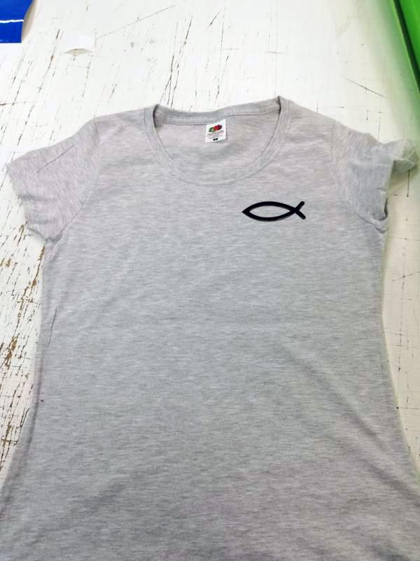 Szara koszulka z nadrukowanym czarnym symbolem ryby na piersi, leżąca na białym, lekko zabrudzonym stole.