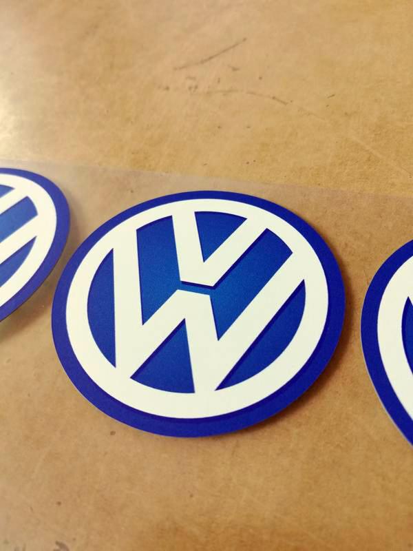 Niebiesko-białe okrągłe emblematy z logo VW na przezroczystej folii, ułożone obok siebie na brązowym tle.