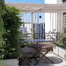 Zaciszny taras w Warszawie z drewnianą pergolą, zielenią w donicach, okrągłym stolikiem i krzesłami składanymi, tworzący kameralną przestrzeń do relaksu.