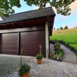 Nowoczesna brama garażowa segmentowa w kolorze ciemnego drewna, z czarnymi prowadnicami, wkomponowana w bryłę domu z kamienną elewacją i brukowanym podjazdem, na tle zielonego wzgórza.