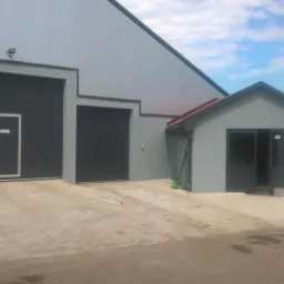 Nowoczesny budynek przemysłowy z trzema czarnymi bramami segmentowymi, szarymi obramowaniami i przylegającym małym budynkiem z drzwiami, świeżo pomalowany, z widocznymi puszkami po farbie na zewnątrz.