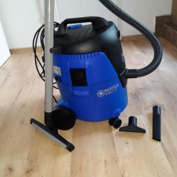 Paromax - Niebiesko-czarny odkurzacz przemysłowy Nilfisk AERO 21 z kompletem ssawek na drewnianej podłodze, widoczne białe drzwi i kartonowe pudło w tle.