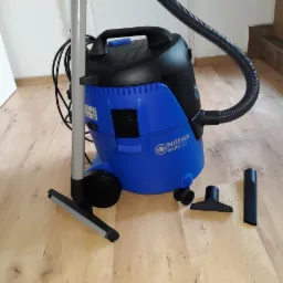 Niebiesko-czarny odkurzacz przemysłowy Nilfisk AERO 21 z kompletem ssawek na drewnianej podłodze, widoczne białe drzwi i kartonowe pudło w tle.