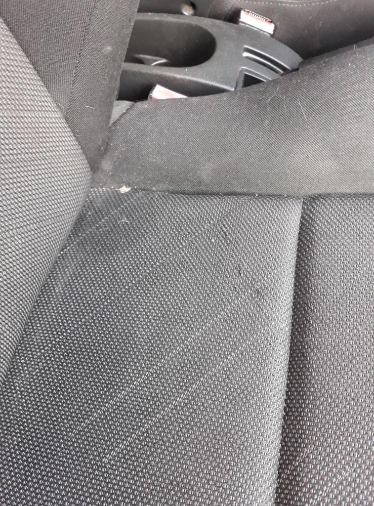 Szare, tekstylne siedzenie samochodowe z widocznymi zabrudzeniami i drobnymi plamami, z fragmentem konsoli środkowej w tle.