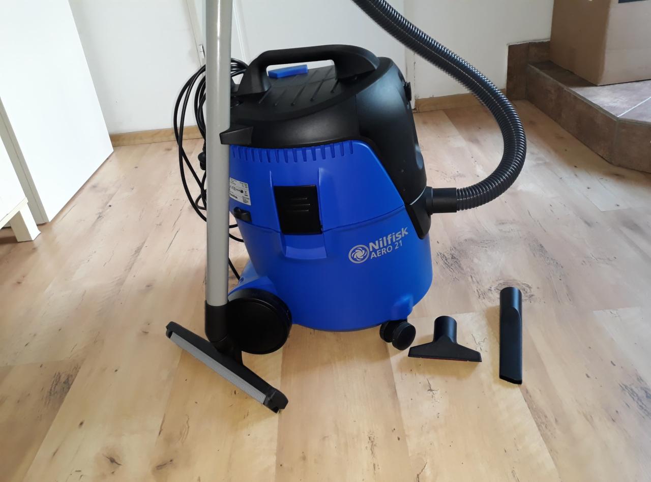 Niebiesko-czarny odkurzacz przemysłowy Nilfisk AERO 21 z kompletem ssawek na drewnianej podłodze, widoczne białe drzwi i kartonowe pudło w tle.