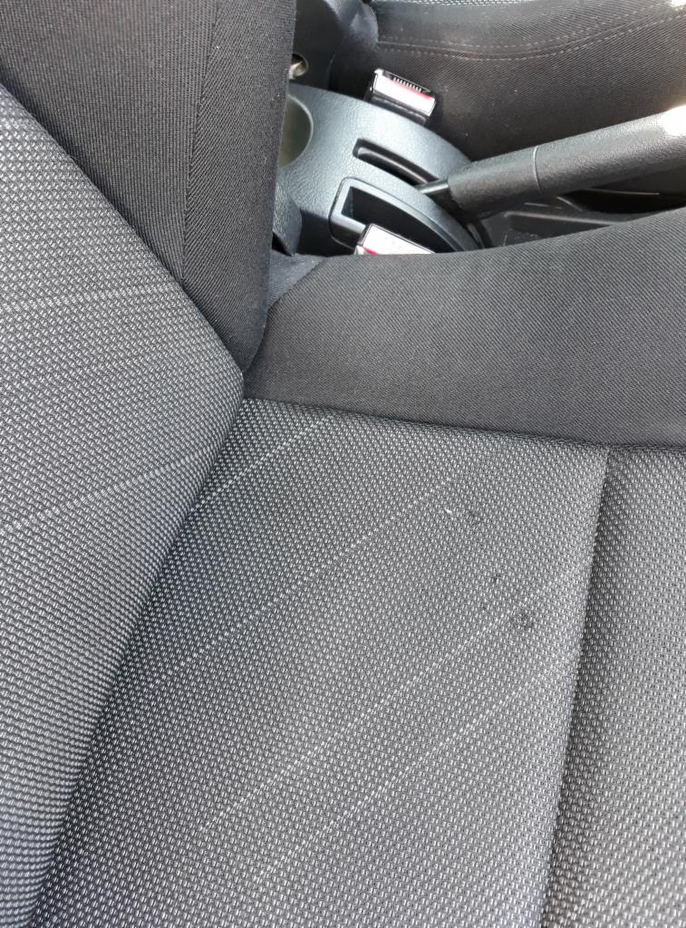 Fragment czarnej, tekstylnej tapicerki samochodowej z widocznymi drobnymi plamami i elementami konsoli środkowej.