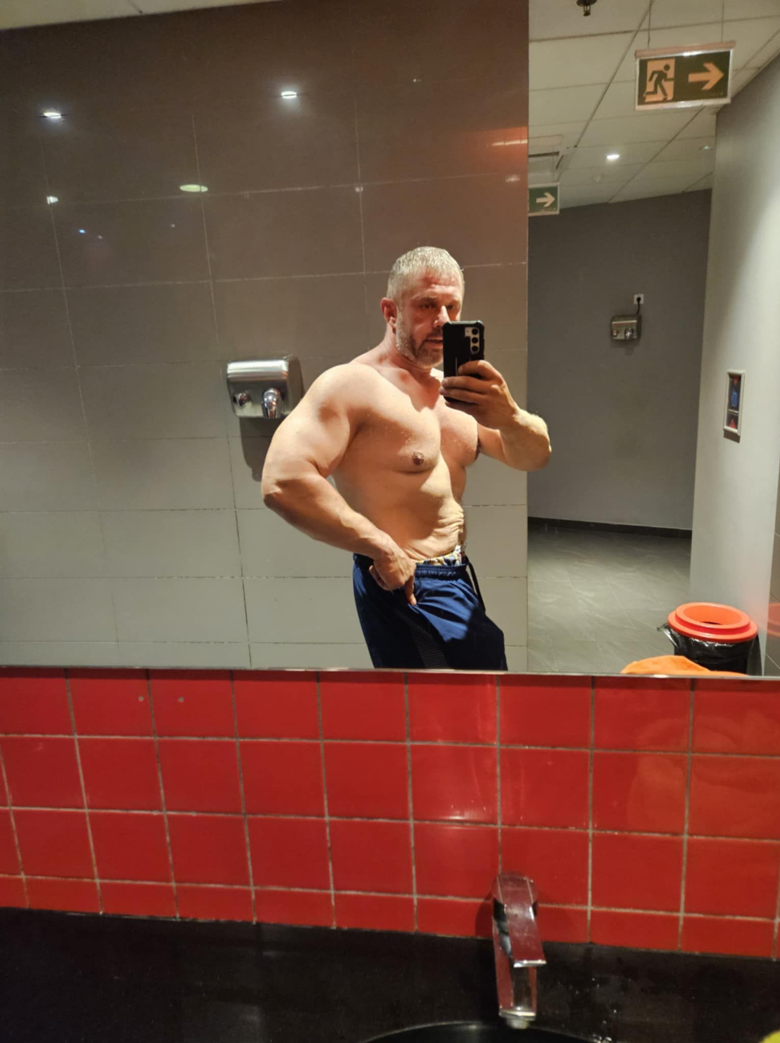 Mężczyzna pozuje, prezentując muskulaturę w lustrze łazienki na siłowni. Widoczny fragment umywalki z czerwonymi kafelkami i znak ewakuacyjny.