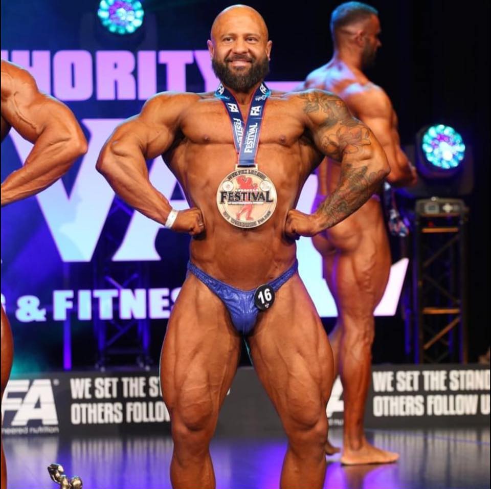Uśmiechnięty kulturysta na scenie, prezentujący muskulaturę, z medalem festiwalowym na szyi i numerem 16 na stroju. W tle inni zawodnicy.