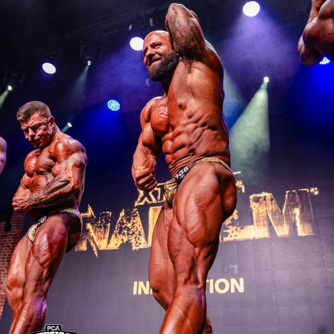 Mężczyzna z numerem 198 prezentuje muskulaturę na scenie podczas zawodów kulturystycznych. Widoczny drugi zawodnik, reflektory i logo imprezy.