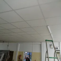 Sufit podwieszany kasetonowy z widocznymi lampami, drabina aluminiowa oparta o ścianę, w tle osoba wchodząca do pomieszczenia.