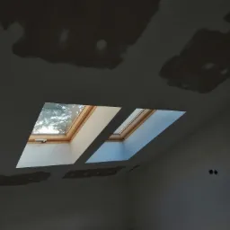 Pokój w trakcie prac wykończeniowych: widoczne dwa okna dachowe, ściany i sufit z widocznymi śladami po szpachlowaniu i przygotowaniu pod malowanie.