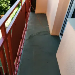 Odświeżony balkon z pomalowaną na czerwono balustradą, szarą podłogą i ścianami w odcieniach beżu, widoczny przewód elektryczny.
