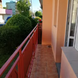 Balkon po remoncie z pomarańczową elewacją, czerwonymi barierkami i brązowymi płytkami podłogowymi, widok na zieleń i budynki w tle.