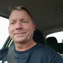 Mężczyzna w samochodzie, zapięty pasami, patrzy w bok z lekkim uśmiechem. Widoczne wnętrze pojazdu i fragment ubrania.