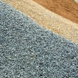 Góra szarego kruszywa o drobnej frakcji, obok hałdy piasku o różnej granulacji i kolorystyce, od beżowego po brązowy, w słoneczny dzień.