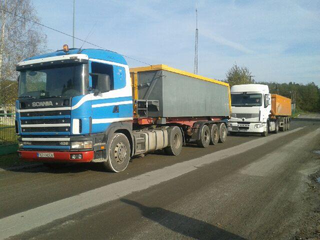 Dwa samochody ciężarowe z naczepami typu wywrotka, pierwszy niebiesko-biały Scania 124L 420 z szarą naczepą z żółtymi krawędziami, drugi biały Renault z pomarańczową naczepą, stojące na asfaltowej...