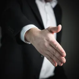 Wyciągnięta dłoń mężczyzny w garniturze, gest powitania lub propozycja zawarcia umowy, zbliżenie na dłoń z widoczną fakturą skóry i detalami mankietu koszuli.