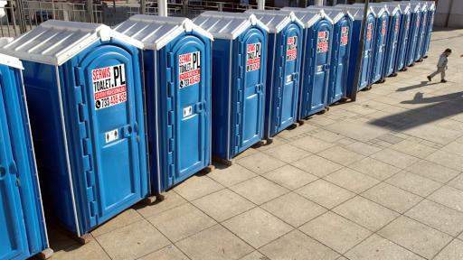 Rząd niebieskich, przenośnych toalet z białymi dachami, ustawionych na placu z betonowej kostki, cień osoby w tle.