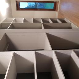 Eurodis Sp.zoo - Wysoka, biała zabudowa regałowa z półkami, widok z dołu, oświetlona przez okno dachowe z drewnianą ramą.