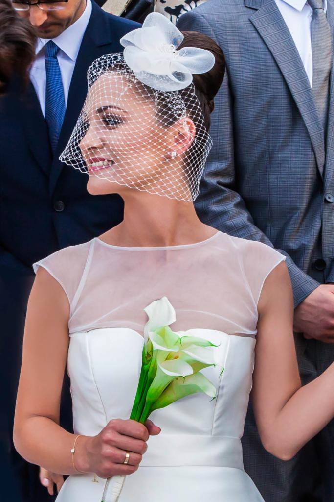 Panna młoda z siateczkowym welonem i kwiatami kalii, uśmiechnięta, w otoczeniu mężczyzn w garniturach.