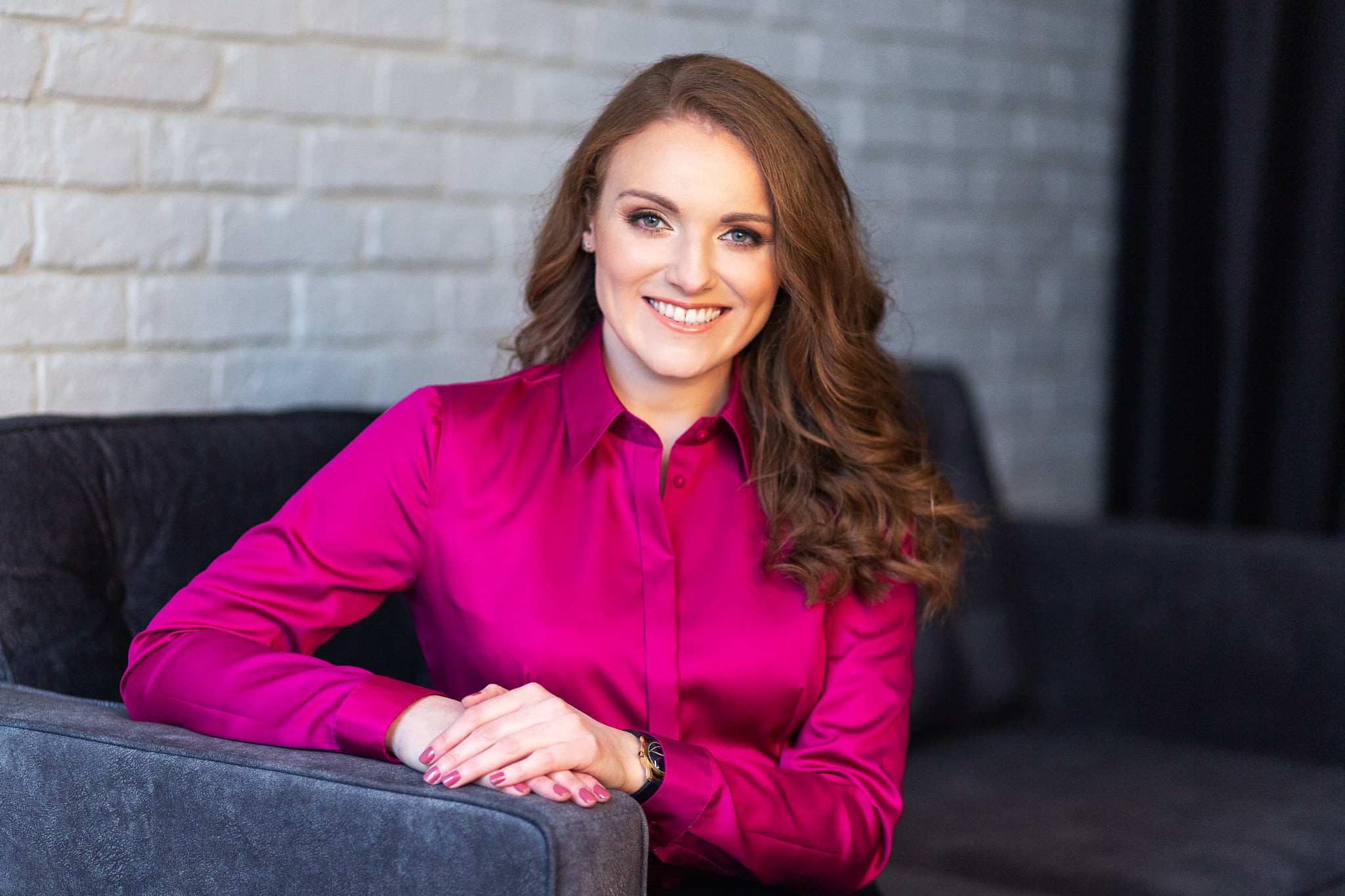 Uśmiechnięta kobieta z profesjonalnym upiększeniem, w fuksjowej satynowej koszuli, siedząca na szarej sofie na tle białej ceglanej ściany i czarnej zasłony.