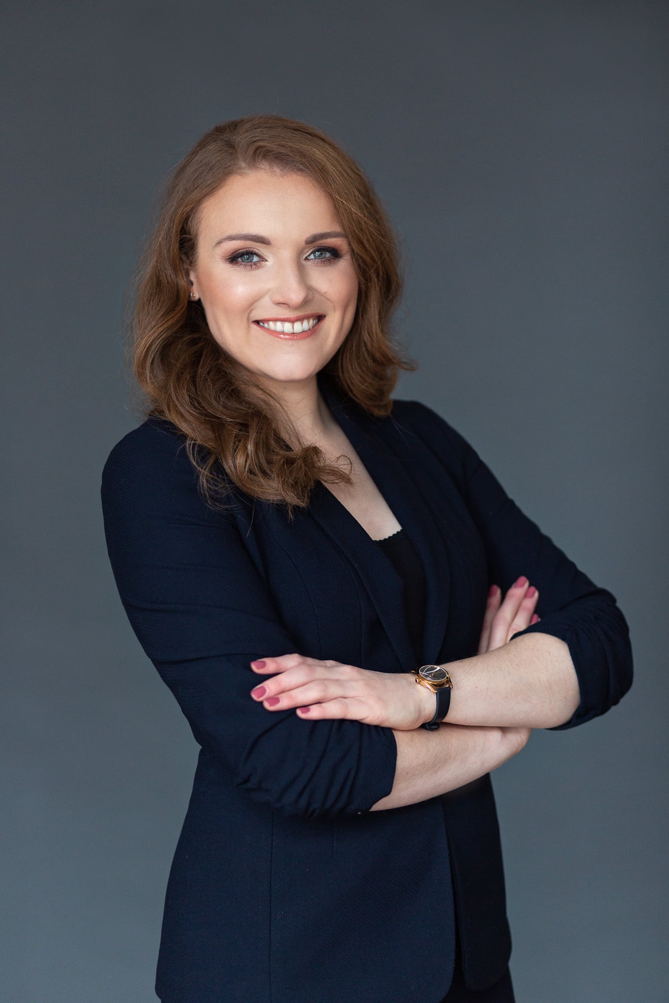 Portret uśmiechniętej kobiety z profesjonalnym, subtelnym upiększeniem, pozującej na tle szarego studia w granatowej marynarce.