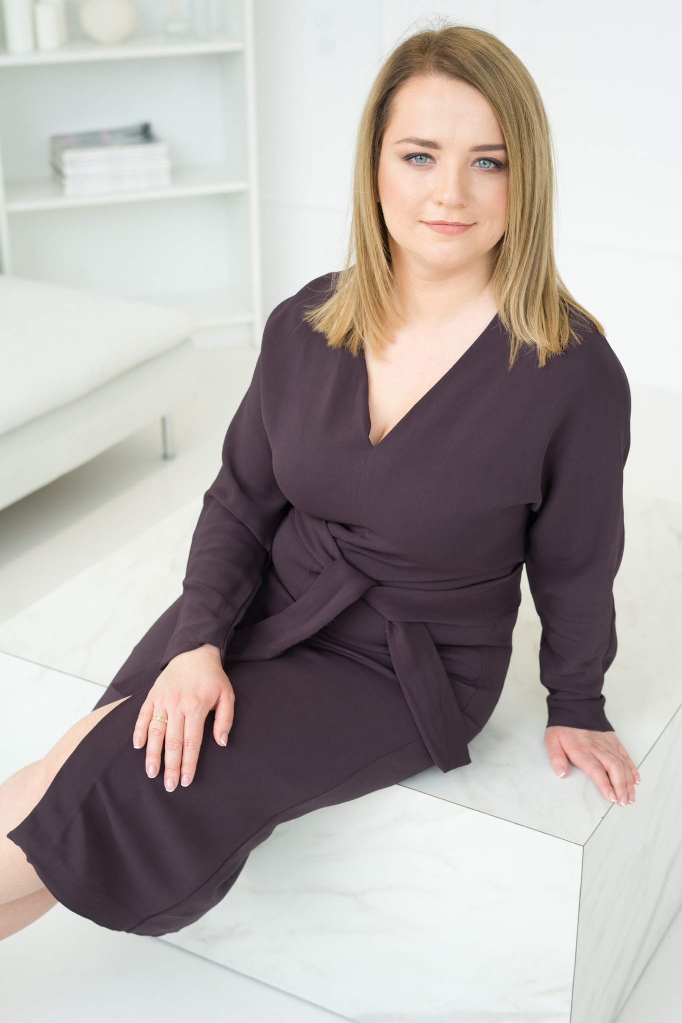 Portret kobiety z blond włosami i niebieskimi oczami, w ciemnej sukience z wiązaniem w talii, siedzącej na białym blacie.