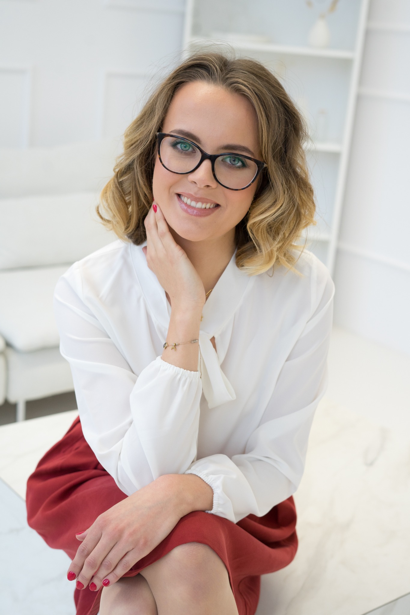 Portret uśmiechniętej kobiety w okularach, z delikatnym, naturalnym podkreśleniem ust i policzków, z widocznym manicure w kolorze czerwonym.