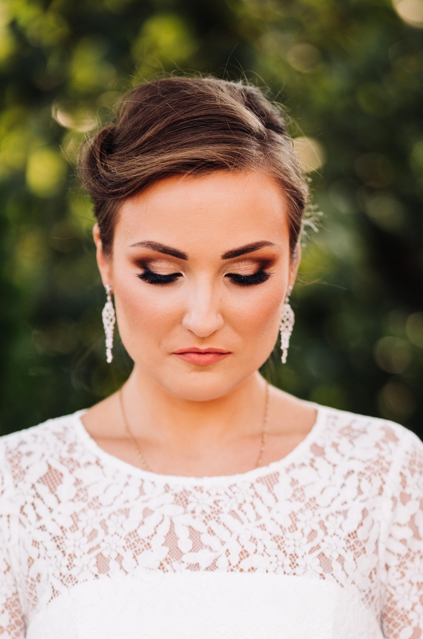 Portret kobiety z zamkniętymi oczami, w makijażu ślubnym z błyszczącymi cieniami, długimi rzęsami i koronkową sukienką, uzupełnionym o wiszące kolczyki.