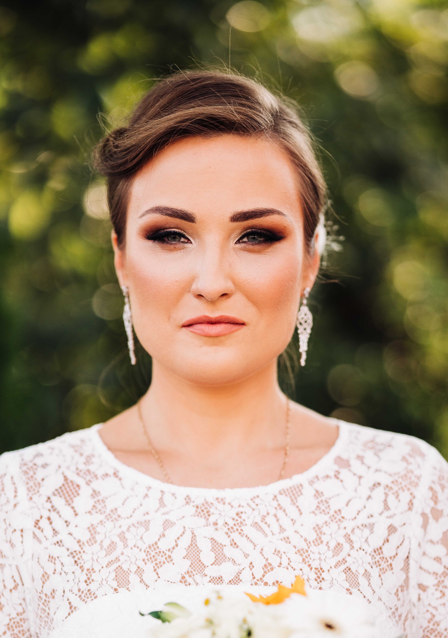 Portret kobiety z upiętymi włosami, w białej koronkowej sukience, z delikatnymi kolczykami i profesjonalnie podkreślonymi oczami, na rozmytym zielonym tle.