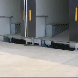 Rampa przeładunkowa z trzema stanowiskami, czarne odbojniki z żółtymi pasami, widoczne elementy mechaniczne pod platformą.