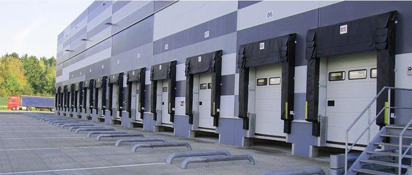 Rząd białych bram przemysłowych z czarnymi uszczelnieniami dokowymi w hali magazynowej, widoczny czerwony samochód ciężarowy w oddali.