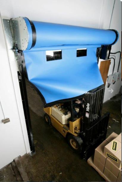 Żółty wózek widłowy Yale przejeżdża przez częściowo otwartą, automatyczną bramę przemysłową z niebieskiego materiału z dwoma okienkami, zamontowaną w magazynie.