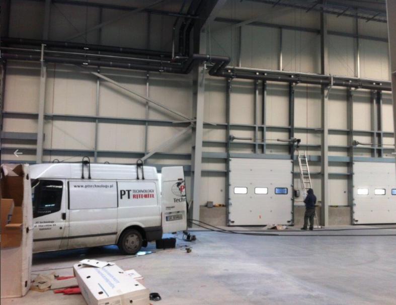 Montaż trzech przemysłowych bram segmentowych w hali, widoczny pracownik na drabinie, biały bus firmy z otwartymi drzwiami i logo parkuje obok, kartony na podłodze.