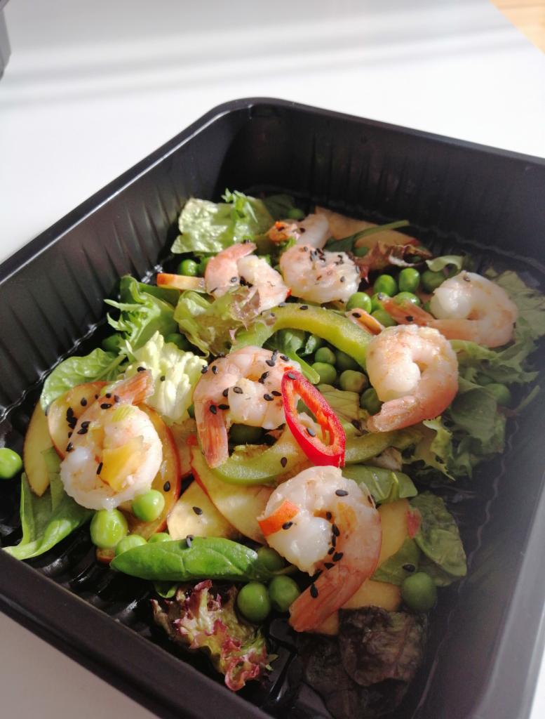 Gotowa sałatka z krewetkami, groszkiem, papryką i jabłkiem, posypana czarnym sezamem, w czarnym pojemniku na białym tle.