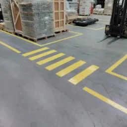 Ż&oacute;łte pasy wyznaczające przejście dla pieszych i strefy bezpieczeństwa na szarej, betonowej podłodze magazynu, z w&oacute;zkiem widłowym i paletami w tle.