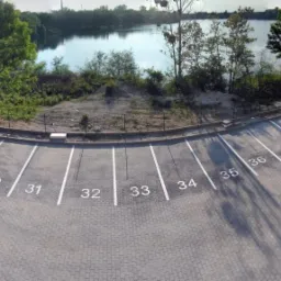 Nowo wybudowany parking z numerowanymi miejscami, widok z góry w słoneczny dzień, w tle zbiornik wodny i zieleń, na parkingu stoi mała koparka.