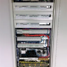 Rozdzielnica elektryczna w metalowej obudowie z widocznymi bezpiecznikami, przekaźnikami i okablowaniem w kolorach niebieskim, czerwonym, żółto-zielonym oraz czarnym, uporządkowane przewody...