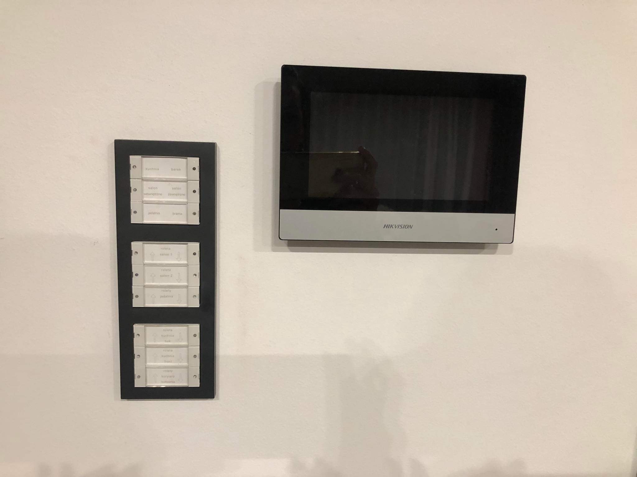 Panel sterowania roletami z opisami pomieszczeń oraz monitor domofonu marki Hikvision zamontowane na białej ścianie.