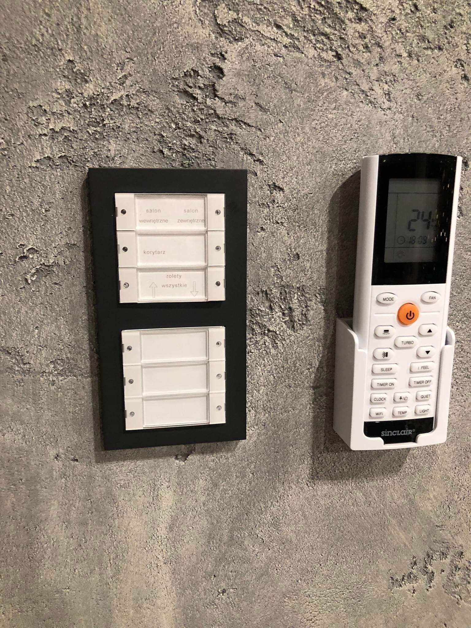 Panel sterowania oświetleniem z opisem stref (salon wewnętrzny/zewnętrzny, korytarz, rolety) oraz pilot do klimatyzacji Sinclair zamontowany na ścianie o szarej, chropowatej fakturze.
