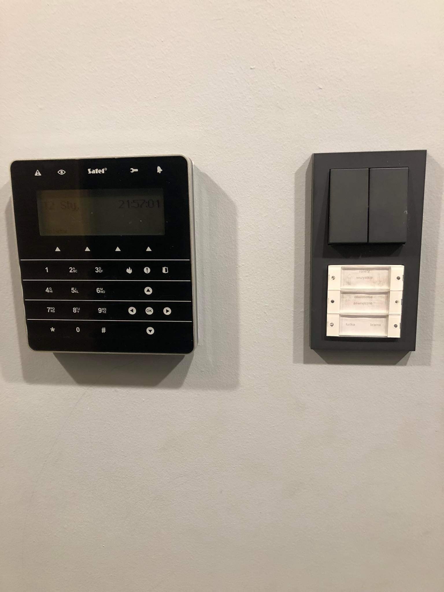 Panel sterowania alarmem Satel z wyświetlaczem LCD i klawiaturą numeryczną, obok czarny panel z włącznikami światła i opisami funkcji (rolety, oświetlenie, furtka/brama) na szarej ścianie.