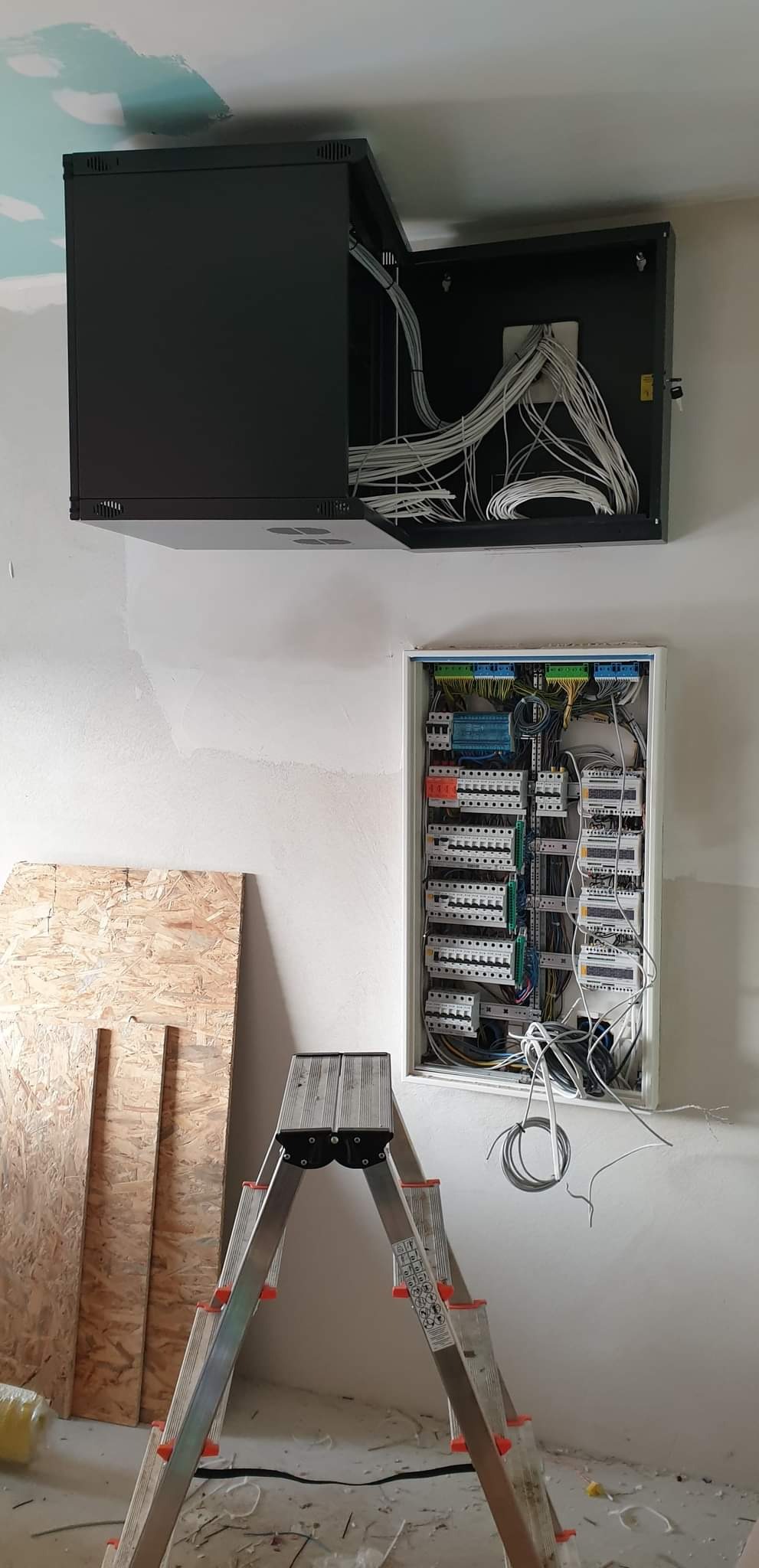 Rozdzielnica elektryczna w trakcie montażu: otwarty czarny panel z kablami u góry, biała skrzynka z bezpiecznikami poniżej, drabina aluminiowa i płyty OSB obok, surowa ściana w tle.
