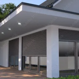 Nowoczesny dom z szarymi roletami zewnętrznymi opuszczonymi na oknach, widok z zewnątrz na taras z drewnianą podłogą, oświetlenie punktowe w podbitce dachu.