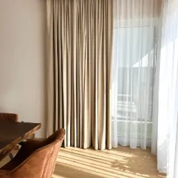 Beżowe, grube zasłony i białe, zwiewne firanki w salonie, widoczne promienie słoneczne na podłodze, stół i krzesło.