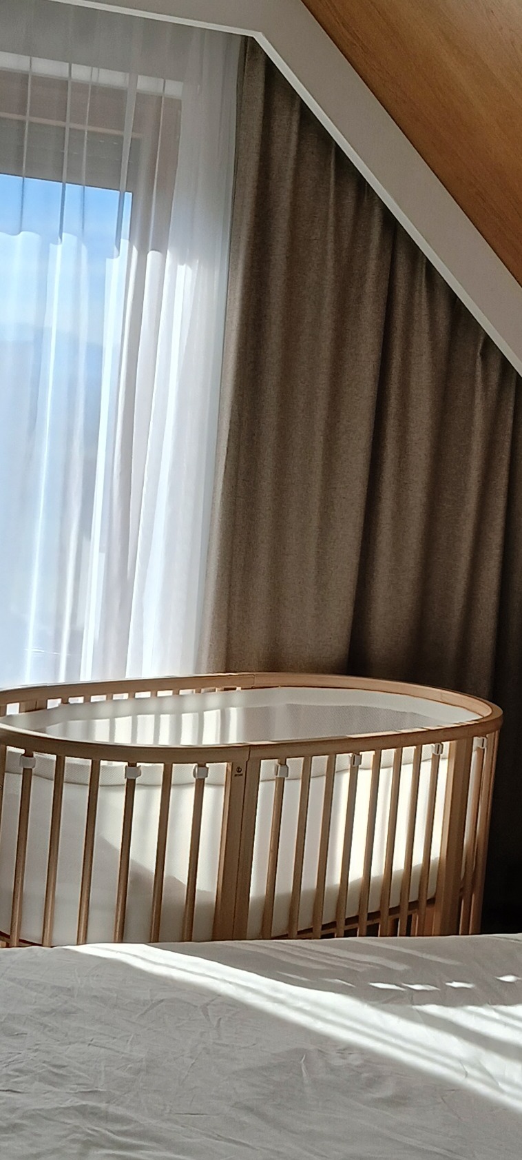 Owalne łóżeczko dziecięce z jasnego drewna, ustawione przy oknie z białymi, przejrzystymi firankami i brązowymi zasłonami, w pokoju ze skośnym sufitem.