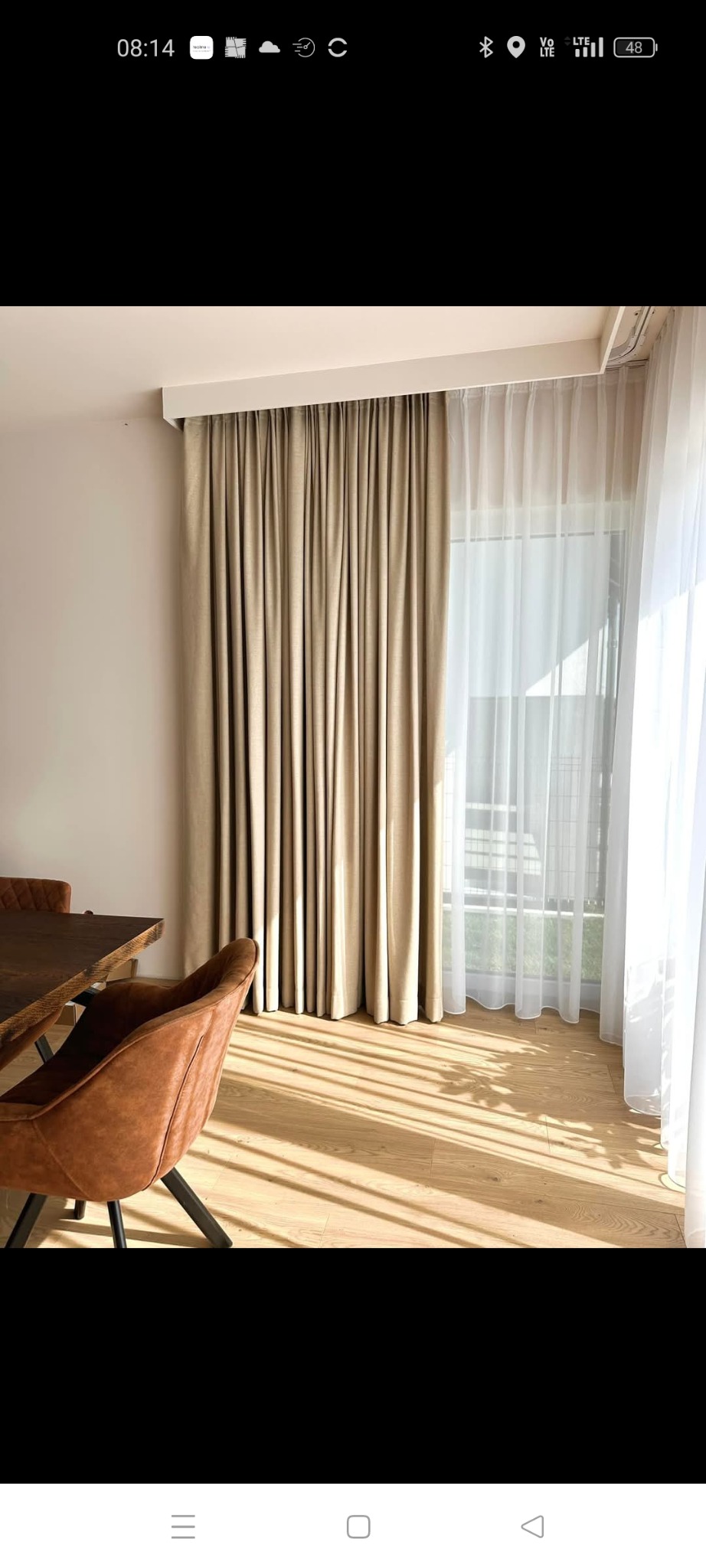 Beżowe, grube zasłony i białe, zwiewne firanki w salonie, widoczne promienie słoneczne na podłodze, stół i krzesło.