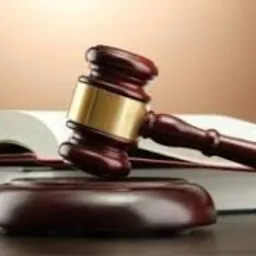 Drewniany młotek sędziowski z mosiężnym pierścieniem, leżący na podstawce, na tle otwartej książki z widocznymi stronami.