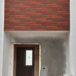 Maniek - Fragment elewacji budynku z wykończeniem z cegły klinkierowej nad otworem drzwiowym w trakcie prac wykończeniowych, widoczne surowe ściany i instalacje.